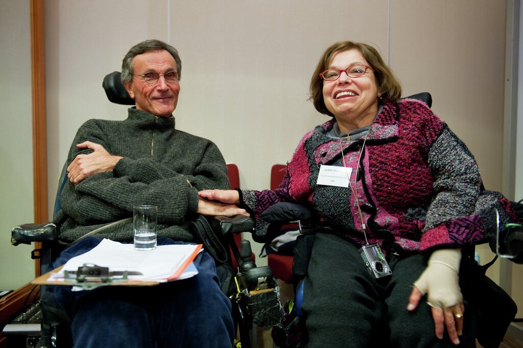 Adolf Ratzka in a gray sweater and with his hands crossed, smiling, next to Judy Heumann. She has a jacket with various shades of pink and grey, red glasses and a white name tag on her chest. She smiles.
