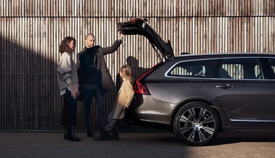 En man öppnar bagageluckan på en Volvo V90. En kvinna står bredvid mannen och ett barn står lutad mot bilen och tittar på de bägge vuxna.
