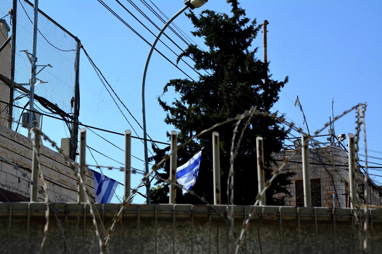 Bildet viser en høy mur med piggtråd på toppen. Bak muren vaier det israelske flagget.