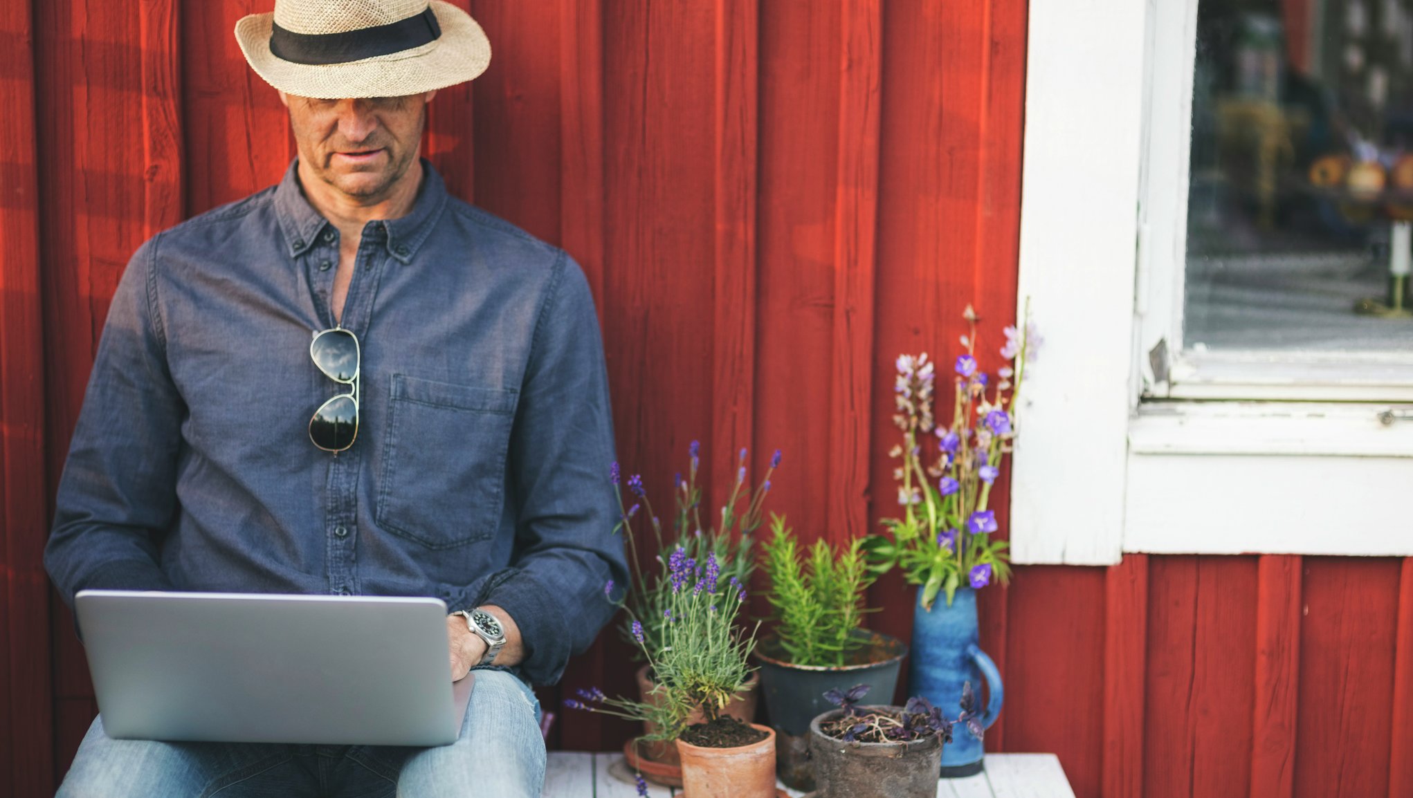 Människa i hatt och med laptop i knäet framför en stuga