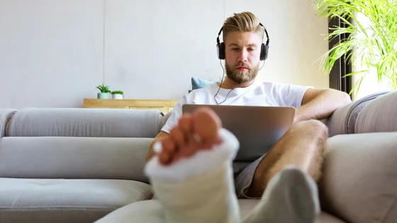 Mies istuu sohvalla tietokoneen ääressä jalka kipsattuna