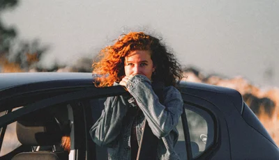 Kvinna med rött hår står lutad mot bildörr.