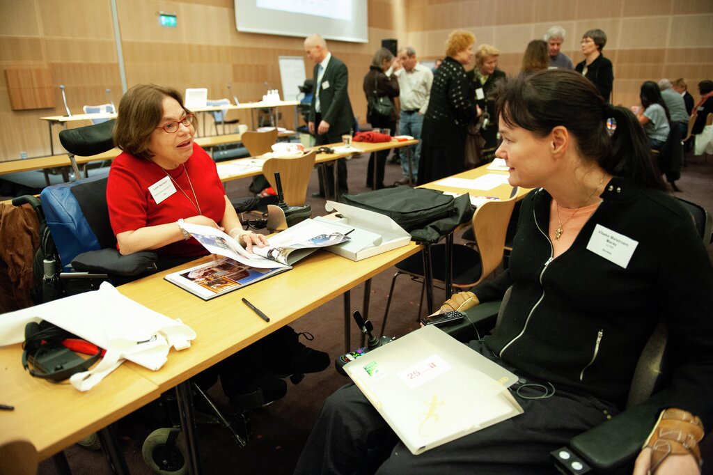 The picture shows Judy Heumann in a red top and Uloba founder Vibeke Marøy Melstrøm in a black jacket and pink top. They sit on opposite sides of a oblong wooden conference desk. There are paper, technical equipment and other things on the desk, and you can see many people in the background. This is probably during a break in a seminar or conference.