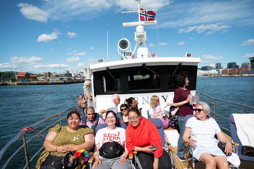 Vi ser en samling av elleve mennesker på båten Arnøy. Det er sol og de er på tur i Oslofjorden. Alle smiler til kameraet og ser ut til å kose seg. Flere har på seg t-skjorter der det står "Stolt, sterk, synlig!"