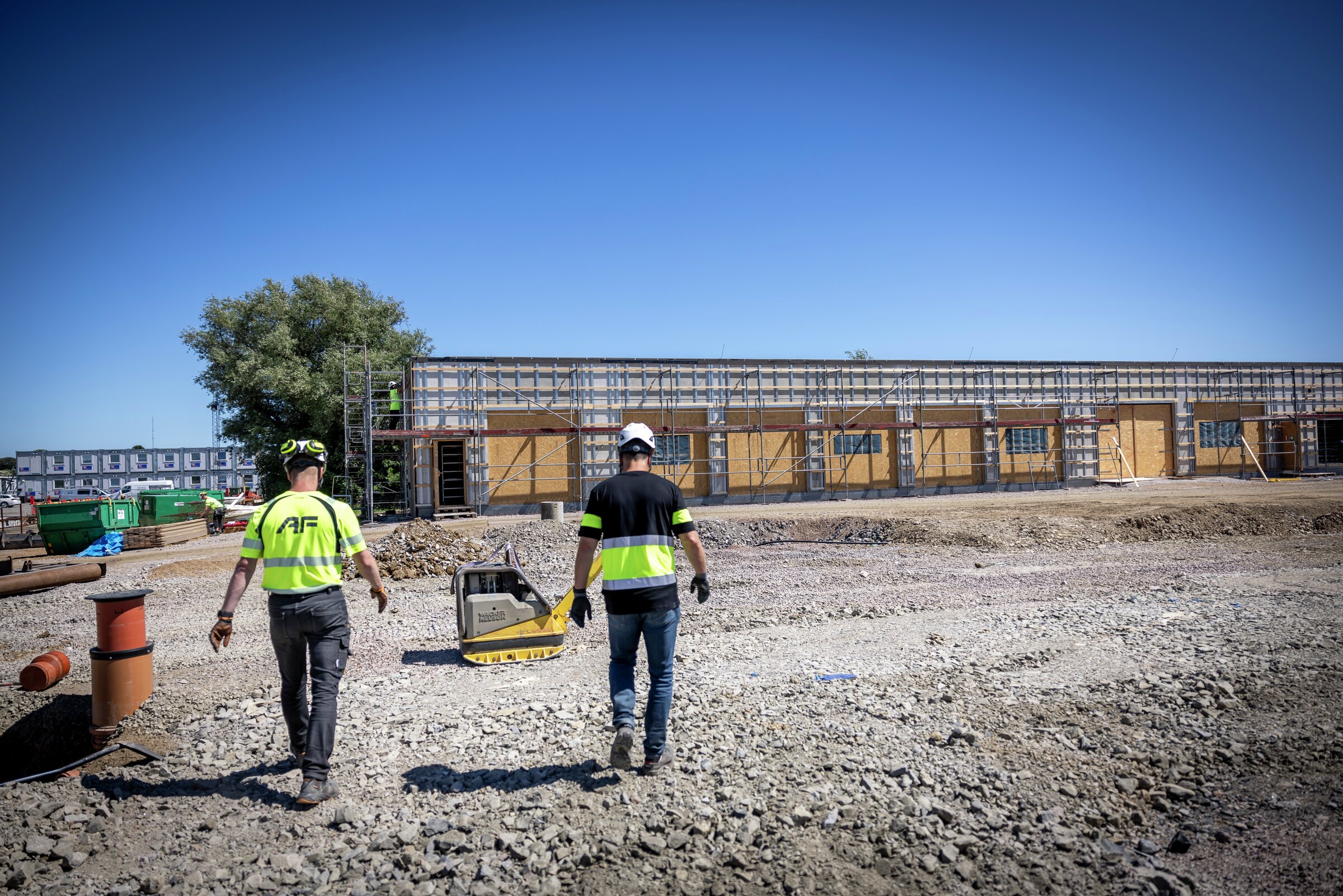 Två byggnadsarbetare i reflexvästar går mot en byggnad under uppförande på en grustomt.