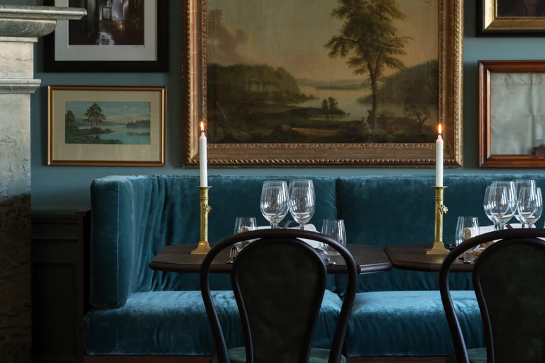 Elegant restauranginredning med blå sammetssittplatser, bord med levande ljus och landskapsmålningar som pryder mörkblå väggar.