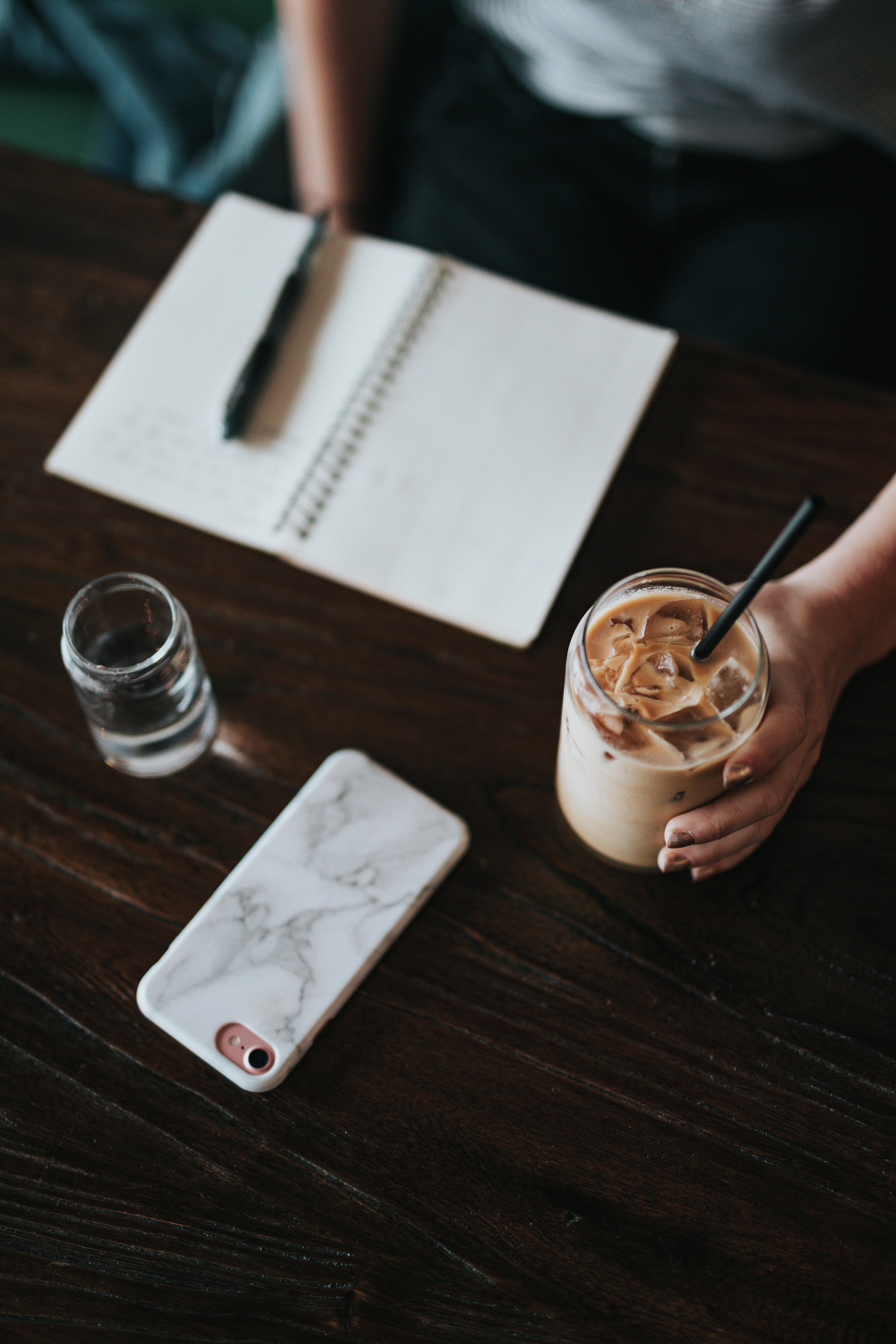 Et bord med en notatblokk, en penn, et vannglass, en iskaffe og en smarttelefon på. Det sitter en person ved bordet.
