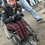 Helle-Viv sitter i rullestolen med skitne sko. I støvet på skoene står det "Fuck off".