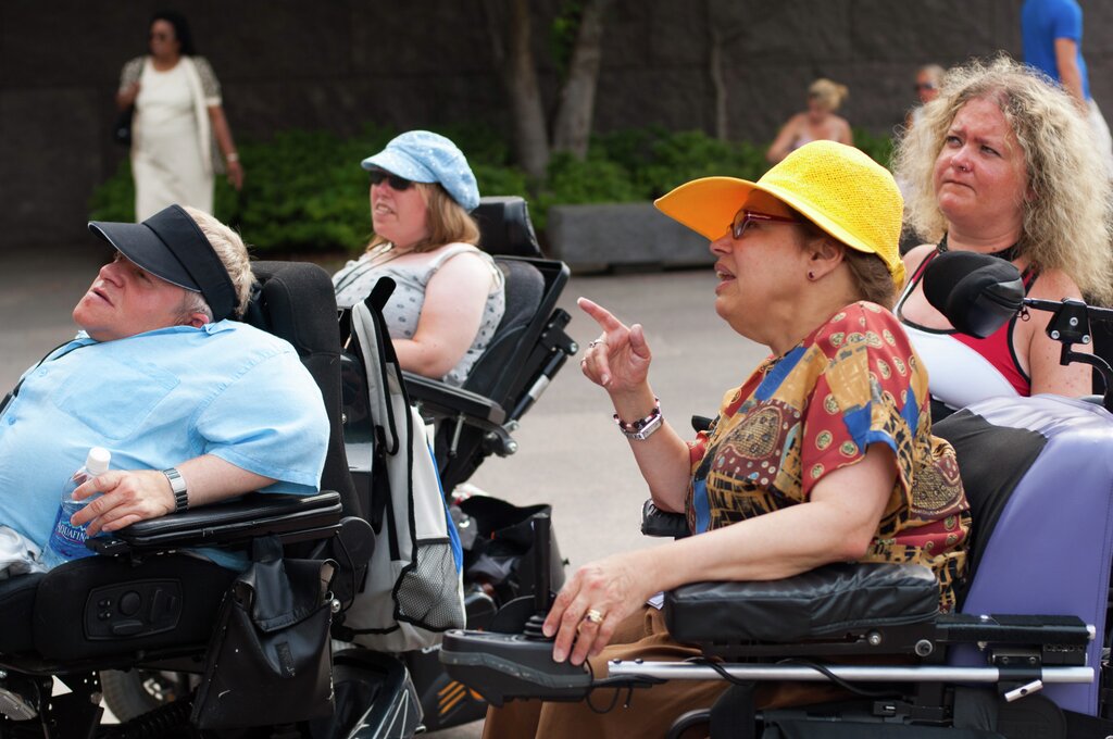 Judy Heumann in conversation with, among others, Knut Flaaum, next to the statue of former president Franklin D. Roosevelt. Heumann wears a yellow sun hat, a blouse in many different colours, and is talking to Knut Flaaum, in a light blue shirt and black parasol.