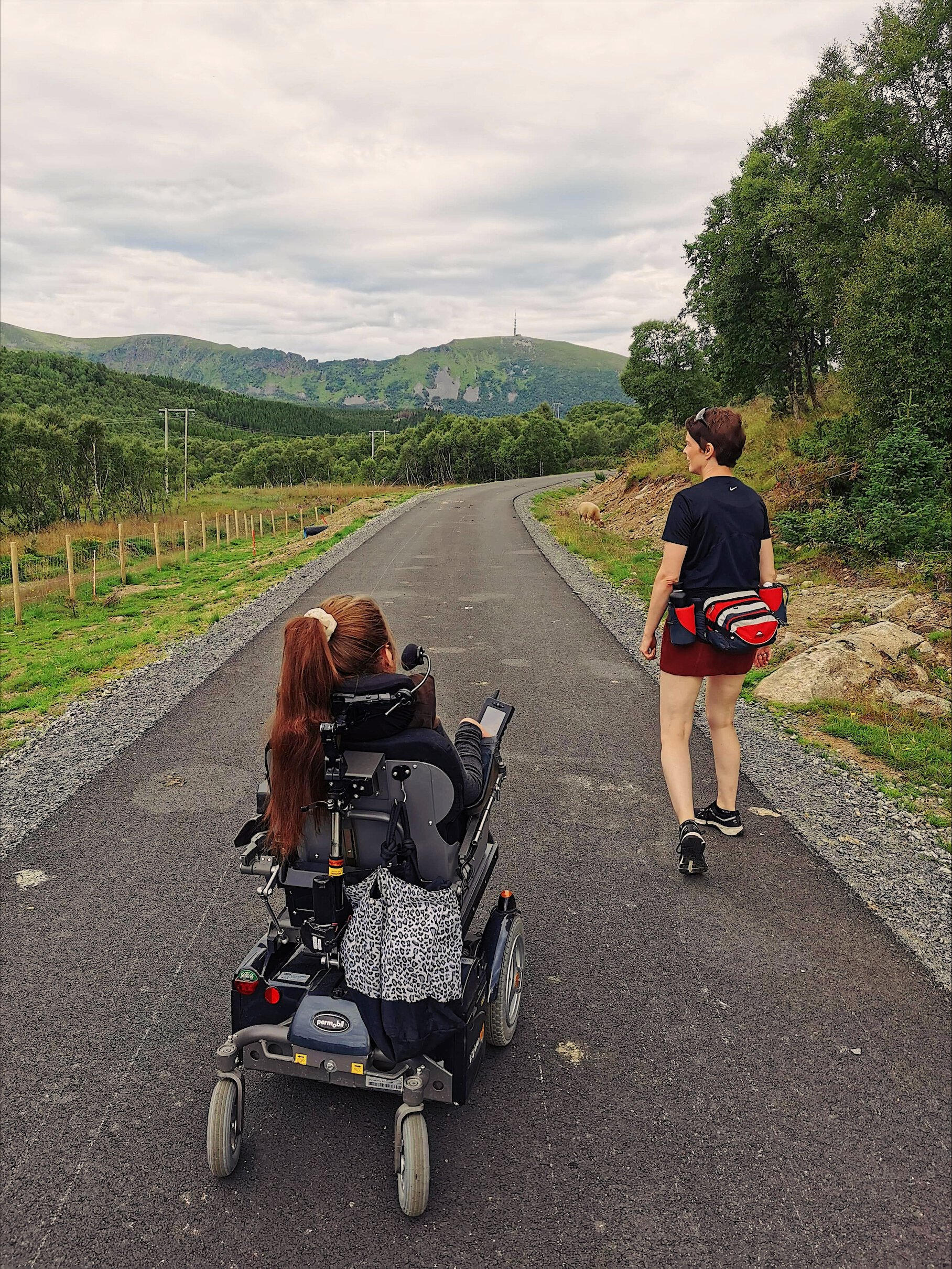 Ingeborg og hennes venninne på tur, veien er asfaltert. Det er fin natur, gresset er grønt, og fjellet kan skimtes i bakgrunn. 