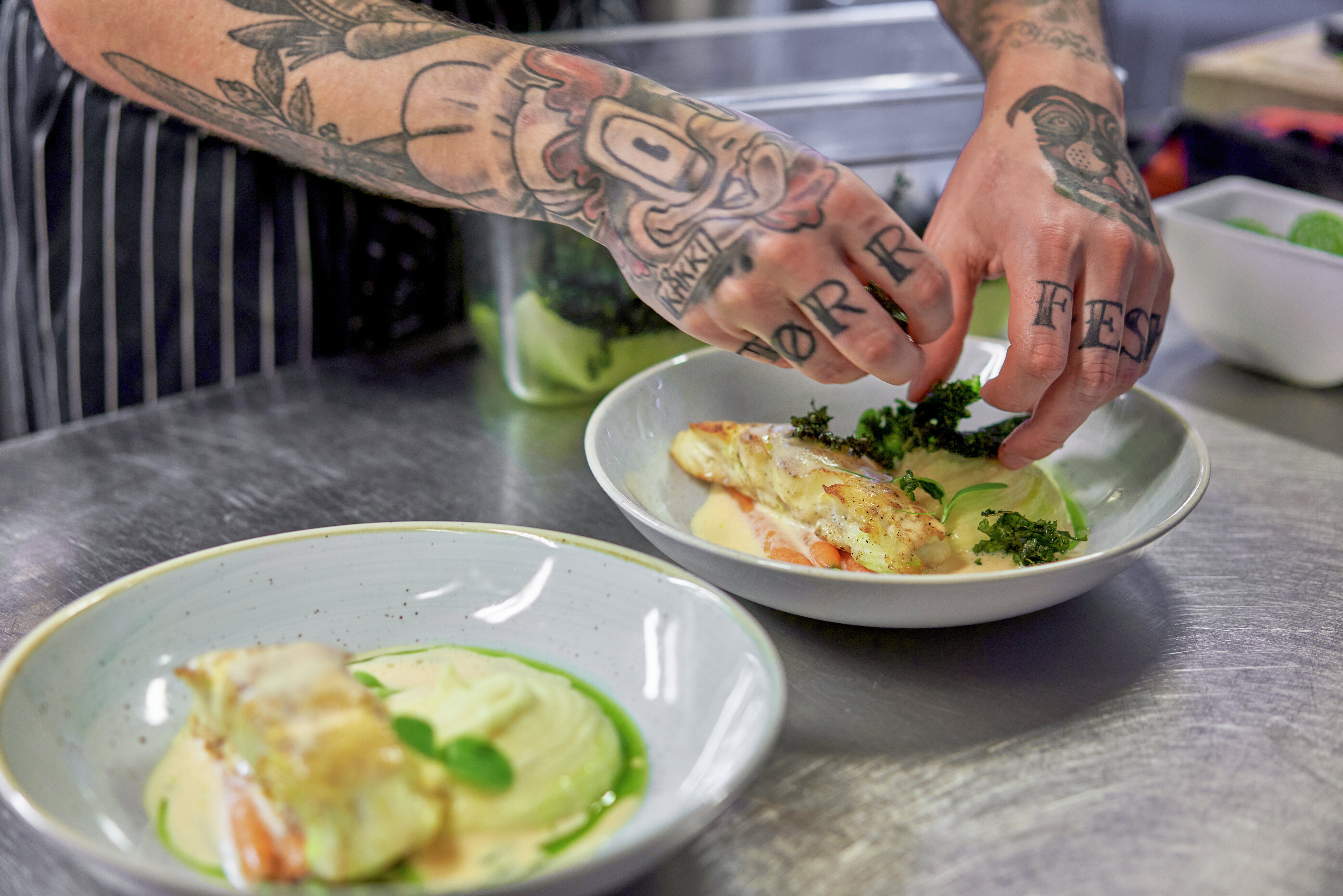 Two hands with tattoes are plating a fish dish in a deep plate.