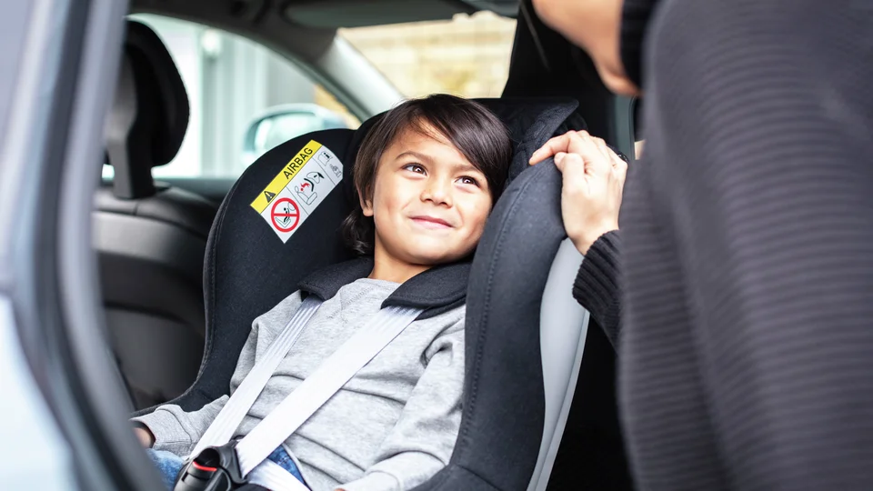 Pojke fastspänd i bakåtvänd bilbarnstol tittar upp mot föräldrar som står i bildörren.