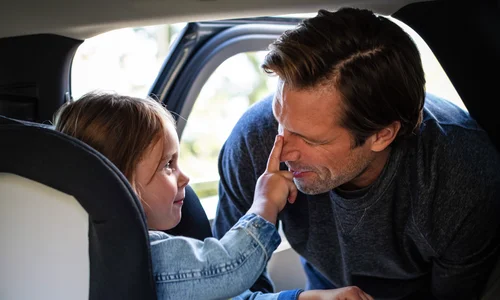 Flicka i bakåtvänd bilbarnstol petar sin pappa på näsan.