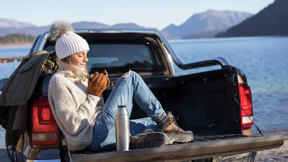 Prie ežero pastatytas pikapas, o jame sėdinti moteris ramiai geria kavą. 