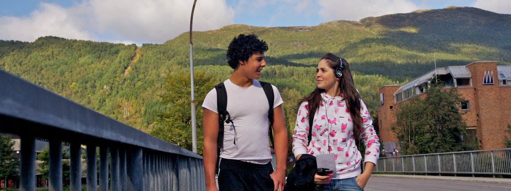 Foto av to ungdomar, ei jente og ein gut, gåande over ei bru i Førde. Dei snakkar saman, ho har øyretelefonar på og han ber ei brusflaske. 